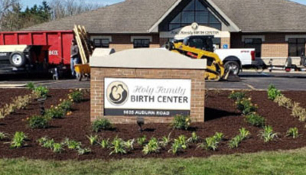 Exterior view of Holy Family Birth Center with a landscaped sign reading 'Holy Family Birth Center' in front; construction vehicles are in the background.