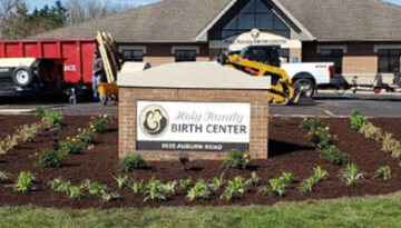 Exterior view of Holy Family Birth Center with a landscaped sign reading 'Holy Family Birth Center' in front; construction vehicles are in the background.