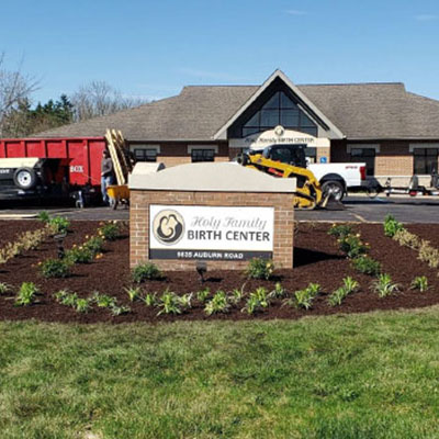 Exterior view of Holy Family Birth Center with a landscaped sign reading 'Holy Family Birth Center' in front; construction vehicles are in the background.