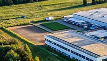 Aerial view of a white, flat-roofed industrial building with parking and green fields surrounding it, bordered by trees and hedges.