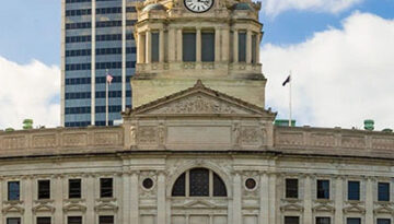 Front view of a grand city hall with a clock tower and dome, with a modern skyscraper behind and a blue sky above