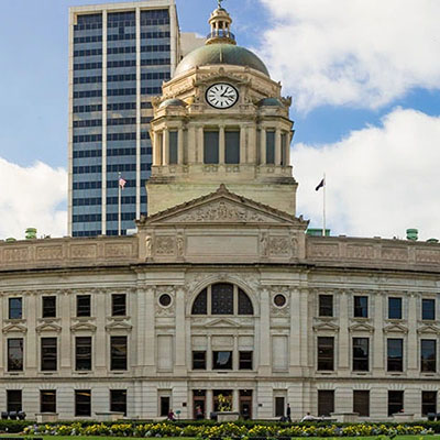 Front view of a grand city hall with a clock tower and dome, with a modern skyscraper behind and a blue sky above